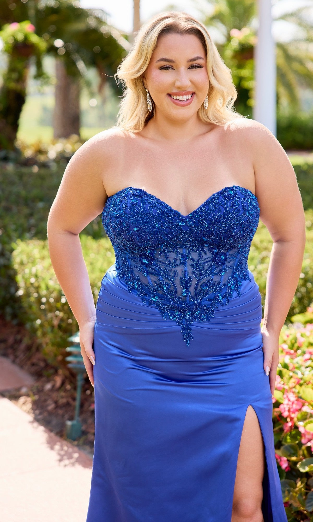 A woman wearing the Primavera 14109 long plus-size prom dress with a strapless satin sheath skirt and sheer embroidered bodice smiles outdoors at the camera.